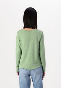 Maglietta verde a maniche lunghe realizzata in tessuto morbido, con scollo rotondo, dettaglio della cucitura posteriore e orlo leggermente curvo. Abbinata a jeans azzurri chiari.