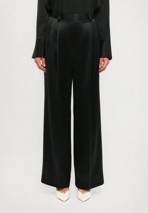 Black high-waist wide-leg pants with front pleats, worn with a black top and white pointed-toe heels on a white background.