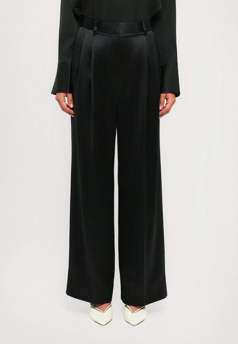 Black high-waist wide-leg pants with front pleats, worn with a black top and white pointed-toe heels on a white background.
