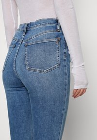 Jeans em denim de lavagem escura com uma cintura média, apresentam bolsos traseiros e detalhes de costura subtil. O tecido tem um acabamento suave, ligeiramente texturizado.