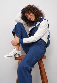 Femme souriante aux cheveux bouclés portant une salopette en denim et des baskets blanches, assise sur un tabouret en bois contre un mur blanc, posant sa main sur sa tête.