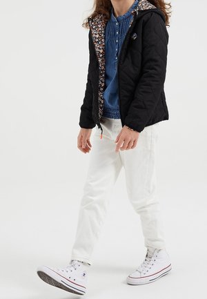 Lapsi kävelee päällään musta tikattu takki, jossa on kukkakuosinen vuori, sininen paita, valkoiset housut ja valkoiset korkeavartiset tennarit.