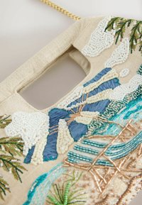 Sac à main en tissu beige, avec une poignée découpée rectangulaire, orné d'une broderie détaillée en perles et fils représentant un paysage avec des arbres, une rivière et un pont.