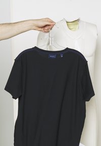 Main tenant un cintre transparent avec un t-shirt noir GANT devant un torse de mannequin blanc sans tête sur un fond uni.