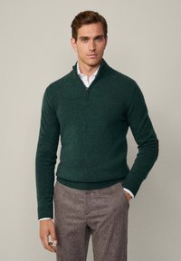 Dunkelgrüner Zip-Pullover aus weichem Strickmaterial, mit gerippten Bündchen und Saum, getragen über einem weißen Hemd mit Kragen.