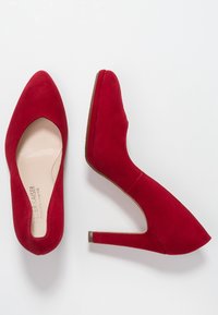 Røde ruskinds høje hæle med spids tå, der har et udskæringsdesign på siderne og en slank stiletto-hæl. Glat indvendig foring.