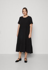 Crna haljina s volanima i kratkim rukavima, okruglim izrezom i lepršavim materijalom. Uparena s sjajnim crnim Mary Jane cipelama. Jednostavna pozadina.