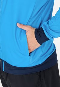 Ljusblå sportjacka med en slät textur och en marinblå kant vid fållen, med en hand i fickan som visar dess funktionella design.