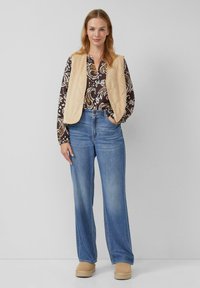 Lichtbeige gewatteerde vest over een bruin patroon blouse, gecombineerd met losse blauwe jeans en tan platform schoenen. Eenvoudig ontwerp, casual stijl.