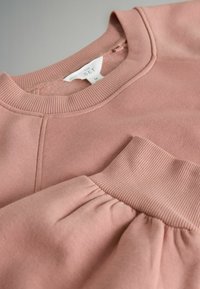 Bluza roz deschis cu guler și manșete cu striații, textură netedă și detaliu adunat la manșete și cusăturile umerilor.