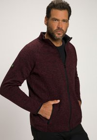 Giacca zip-up in tessuto mélange rosso e nero con colletto alto, due tasche laterali e polsini neri a contrasto. Presenta un piccolo logo verde sulla manica.