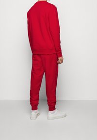 Homme debout, de dos, portant un sweat-shirt rouge et un pantalon de jogging assorti, avec des chaussettes blanches et des baskets blanches, devant un mur uni.