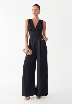 Kobieta w czarnym kombinezonie bez rękawów o szerokich nogawkach, trzymająca beżową kopertówkę, nosząca złote kolczyki, bransoletkę i sandały.