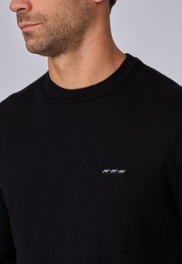 Pull noir en maille avec col rond, agrémenté de petites vaches blanches brodées sur la zone du côté gauche de la poitrine. Texture douce et design ajusté.