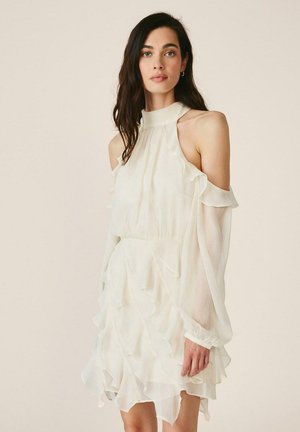 Mujer con cabello largo y oscuro que lleva un vestido blanco con hombros descubiertos, volantes y un escote alto contra un fondo beige liso.