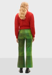 Pull en tricot rouge avec des manches bouffantes, associé à un pantalon à carreaux verts évasés. Des chaussures noires épaisses complètent la tenue.