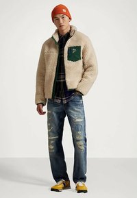 Νεαρός άνδρας φορεί ένα μπεζ σακάκι sherpa, πορτοκαλί πλεκτό σκουφάκι, καρό πουκάμισο, φθαρμένα τζιν και κίτρινα αθλητικά παπούτσια, στέκεται μπροστά από έναν ουδέτερο φόντο.