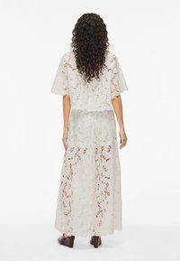 Weißes Spitzenkleid mit kurzen Ärmeln, floralem Muster, gestuftem Design, transparenter Textur und gewelltem Saum, kombiniert mit dunkelbraunen Schuhen.