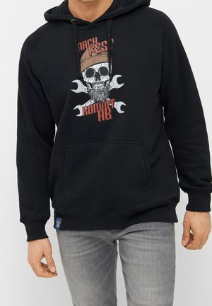 Homme portant un sweat à capuche noir avec un motif de crâne, un bonnet marron et une barbe, des clés croisées derrière le crâne et un texte rouge sur le devant.