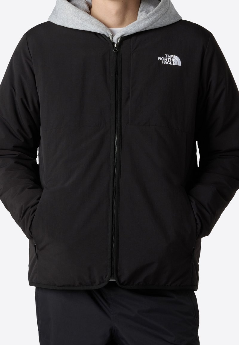 Personne portant une veste zippée noire par-dessus un sweat à capuche gris, les mains dans les poches, avec le logo The North Face sur la poitrine.