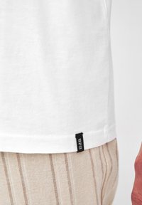 T-shirt en coton blanc avec une étiquette noire marquée "BZB" sur l'ourlet, associé à un tissu rayé beige. Design simple et épuré.