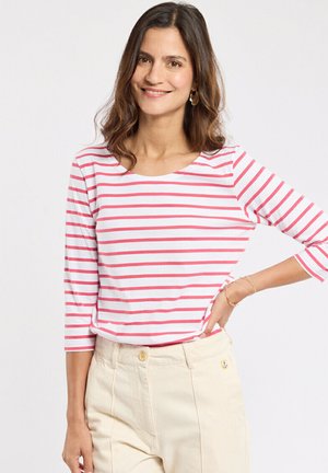 Femme souriante portant un haut à manches trois-quarts blanc et rose à rayures et un pantalon beige taille haute avec des bijoux en or à un poignet.