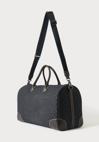 Μαύρη τσάντα duffel με σχέδια, δερμάτινες λαβές, ενισχυμένες γωνίες και ρυθμιζόμενο ιμάντα ώμου σε απλό φόντο.