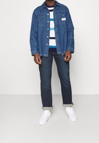 Jeansjacka i medelblått, med två framfickor och silverknappar, bärs över en randig tröja och mörka jeans med uppvikta fållar.