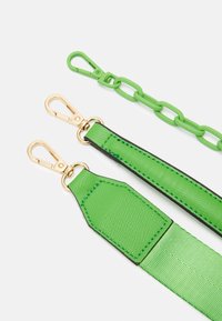 Zelené popruhy na tašku se zlatými kovovými sponami a zeleným plastovým řetězem na bílém pozadí.