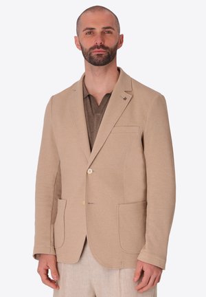Uomo con barba corta che indossa una giacca beige strutturata sopra una camicia marrone e pantaloni chiari su uno sfondo neutro.