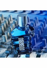 Sininen lasipullo, joka on muotoiltu lihaksikkaaksi ylävartaloksi ja jossa on hopeinen korkki. Etiketissä näkyy selkeästi hopeinen teksti "DIESEL." Taustalla näkyy miksauspöytä.