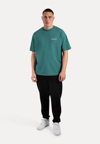 Camiseta de algodón color verde azulado con cuello redondo y mangas cortas, con texto blanco en el pecho, combinada con pantalones sudados negros y zapatillas blancas.