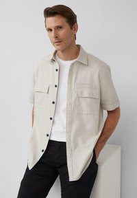 Lichtbeige shirt met korte mouwen, twee borstzakken, marineblauwe knopen en een ontspannen pasvorm, gelaagd over een wit t-shirt en gecombineerd met zwarte broek.