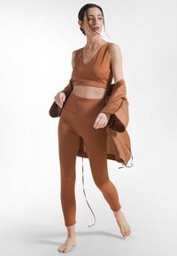 Frau in braunem, ärmellosem Crop-Top und passenden Leggings, barfuß, hält eine locker sitzende braune Jacke und steht vor weißem Hintergrund.