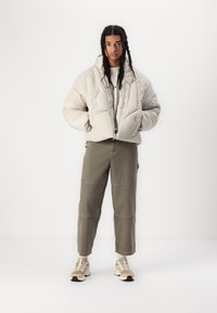 Veste matelassée beige clair avec capuche, texture matelassée, associée à un pantalon large olive et des baskets beiges avec des accents noirs.