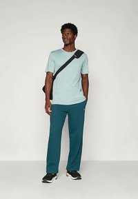 Homem vestindo uma t-shirt azul clara e calças verdes-azulado, de pé com uma mão no bolso, mochila preta cruzada e ténis pretos, contra uma parede simples.