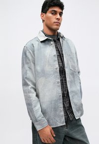 Svetlomodrá denimová bunda s zapínaním na gombíky, dlhými rukávmi a náprsné vrecko, preložená cez čierno-sivú károvanú košeľu.