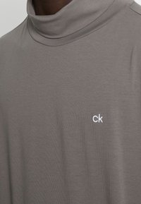 Grauer Rollkragenpullover aus glattem Stoff mit einem kleinen weißen "ck"-Logo auf der linken Seite. Standardpassform mit minimalen Details.