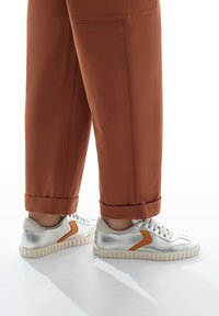 Sneakers argento con finitura texturizzata, caratterizzati da un accento arancione a contrasto e una suola spessa e rigata, abbinati a pantaloni marroni arrotolati.