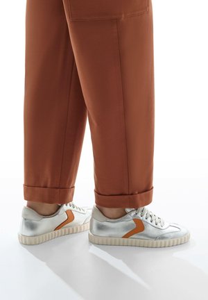 Silver sneakers with a textured finish, featuring a contrasting orange accent and a thick, ridged sole, paired with rolled brown trousers.