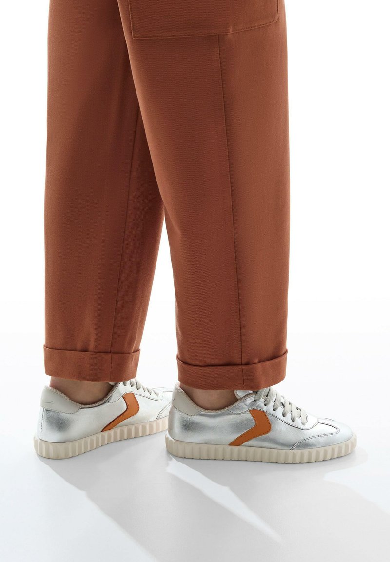 Sneakers argento con finitura texturizzata, caratterizzati da un accento arancione a contrasto e una suola spessa e rigata, abbinati a pantaloni marroni arrotolati.