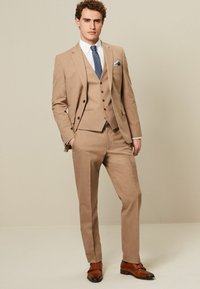 Hellbrauner dreiteiliger Anzug mit Revers, schmal geschnittene Hose und einer blauen Krawatte mit Punkten. Braune Lederschuhe runden das Outfit ab.