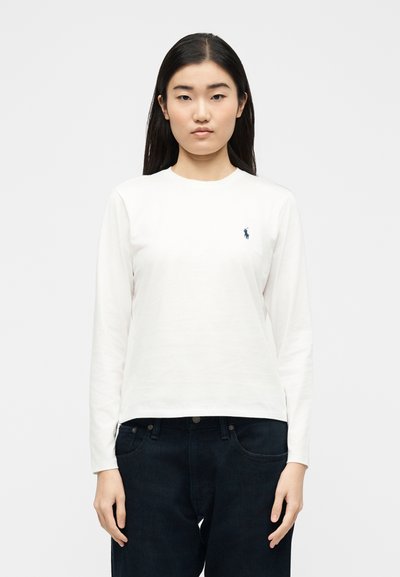 Νεαρή γυναίκα με μακριά σκούρα μαλλιά, φορώντας λευκό πουκάμισο με μακριά μανίκια με λογότυπο σε ναυτικό μπλε και σκούρο τζιν, σε απλό φόντο.