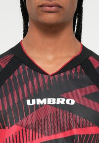 Közeli kép egy személyről, aki fekete és piros mintás sportpólót visel, rajta fehér "UMBRO" felirattal a mellkasán.
