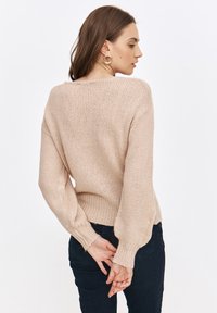 Beżowy sweter wykonany na drutach, o luźnym kroju, z długim rękawem i ściągaczami na mankietach. Ma teksturowany wzór oraz okrągły dekolt. Noszony z ciemnymi dżinsami.