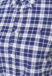 Blå och vit rutig skjorta med knappar gjord av vävt tyg; har en spetsig krage och Polo-logotypen broderad i lila.