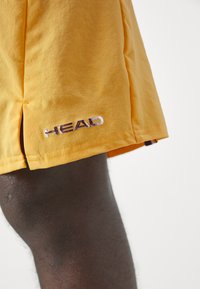 Gelbe Sportshorts aus strukturiertem Stoff, mit einem seitlichen Schlitz und einem kontrastierenden gestickten "HEAD"-Logo in Lila und Weiß.