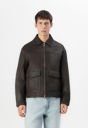 Young man wearing dark brown leather jacket with front zipper and flap pockets, paired with light blue jeans, standing against plain white background.