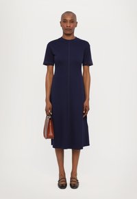 Marineblaues Kurzarmkleid mit geradem Schnitt, das eine Mittelnaht und strukturierte Details aufweist. Kombiniert mit einer kleinen braunen Handtasche und schwarzen Schuhen.