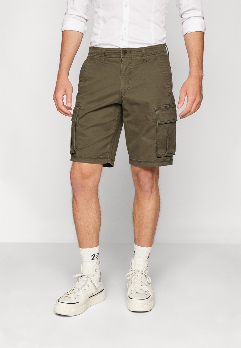 Homme portant un short cargo vert olive, des baskets montantes blanches, des chaussettes blanches et une chemise blanche à manches retroussées.